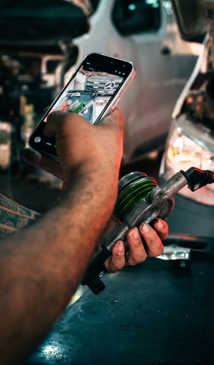 Mecânico da TS Auto Center fotografando a peça com o celular para mandar no WhatsApp do cliente, com uma Sprinter aberta ao fundo